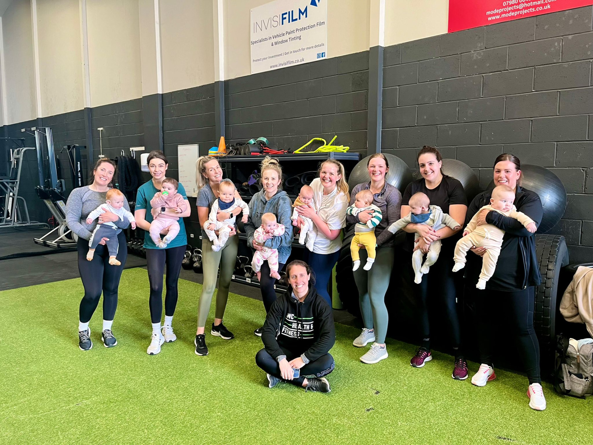 Nine mums standing on the gym's turf area holding their babies, with coach Sally sitting cross-legged in front