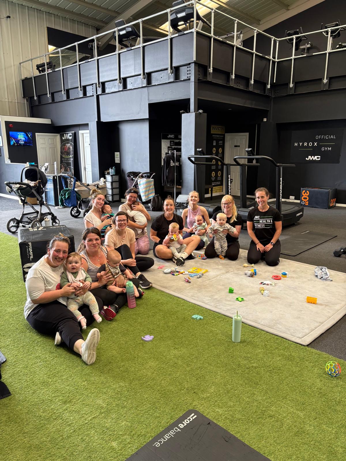 Mums sitting on the turf and a play mat with their babies and toys, with coach Sally kneeling beside them, at JWC Health & Fitness